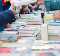 Photograph of stacks of books on tables(no titles visible), with people standing next to the tables.
