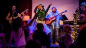 The musicians of Jessa and the Saplings (lead singer, guitar, and banjo) perform onstage in brightly colored costumes