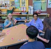 Teens playing a card game around a table with one smiling at the camera Teens playing a card game around a table with one smiling at the camera