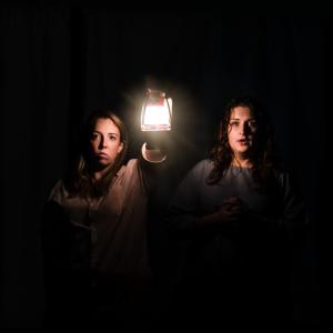 Photo of two opera singers, surrounded by darkness, faces illuminated by the light from a lamp held up by one of them.