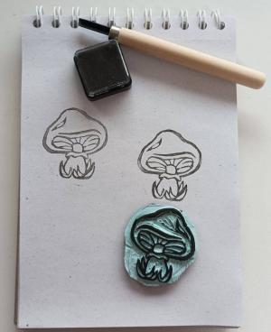 Photo of a rubber stamp carved into the shape of a mushroom, two stamped mushrooms on gray paper, and a carving tool.