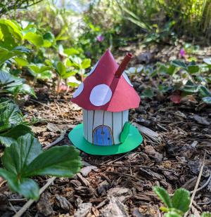 Photo of Gnome Home with white walls, a blue door, and red mushroom cap roof, all made from paper; sitting in a forest.