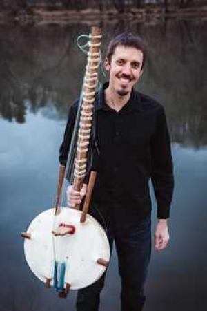 Photo of musician Sean Gaskell, holding a kora (African lute), wearing dark clothing and standing in front of water.