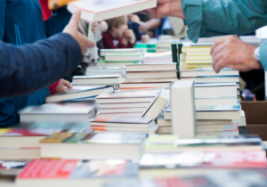 Photograph of stacks of books on tables(no titles visible), with people standing next to the tables.